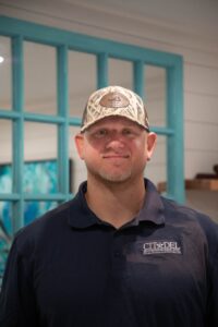 John G, Project Manager and Lead Carpenter at Citadel Enterprises, smiling in a navy shirt with company logo and camo hat, set against a bright blue background.